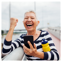 An older woman smiling widely while clenching her fist in happiness, she holds a smartphone towards her, implying she is calling someone
