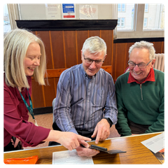 An AbilityNet BT Group Digital Skills Trainer helping two older men with their smart phone